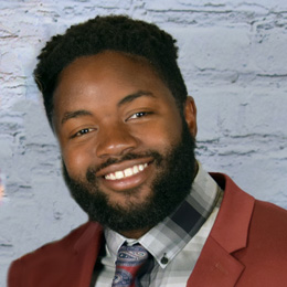 Tarik smiles at the camera sporting short, curly hair and a full beard. He is wearing a maroon jacket over a plaid shirt and a patterned tie.