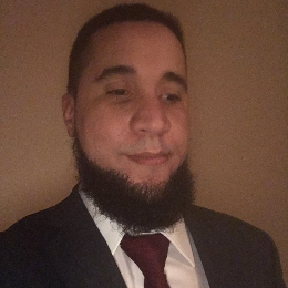 Kris has short dark hair and a full beard. He wears a charcoal gray suit jacket, a white dress shirt, and a burgundy tie.