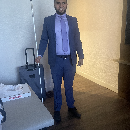 Ammar Tarin is seen wearing a dark blue suit with a light purple shirt and a matching tie. He is holding a white cane in his right hand.