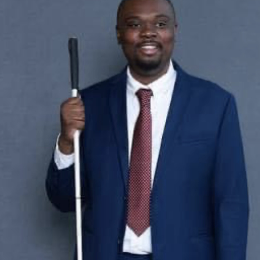 Macaulay Beasley smiling and wearing a dark blue suit, white shirt, and red tie. He is holding a D-Cane in his right hand and is sporting a beard.