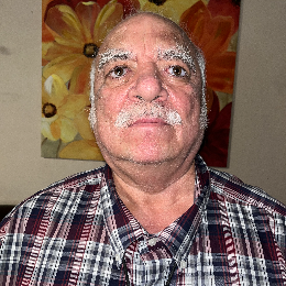 Mark Feliz is pictured with a mustache and short white hair. He is wearing a plaid shirt with a pattern of red, blue, and white colors.