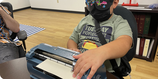 A blind boy reaches forward to check his spelling while working on a Braille writer under learning shades.